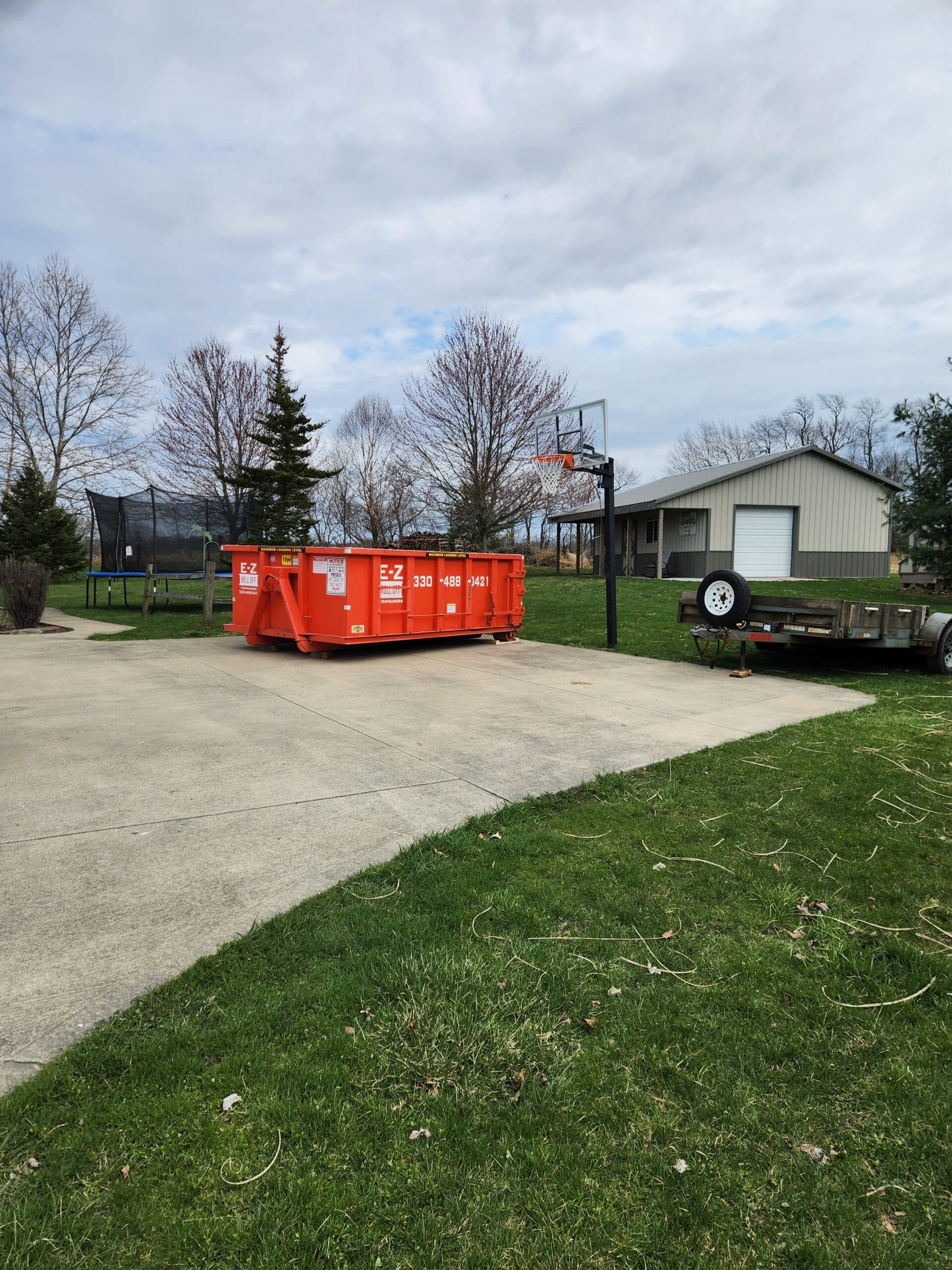 A white truck from E-Z Roll Off Containers with a logo and phone number (330) 488-0421, unloading an orange 10-yard roll-off dumpster on a gravel surface, with a field and trees in the background under a blue sky with clouds.