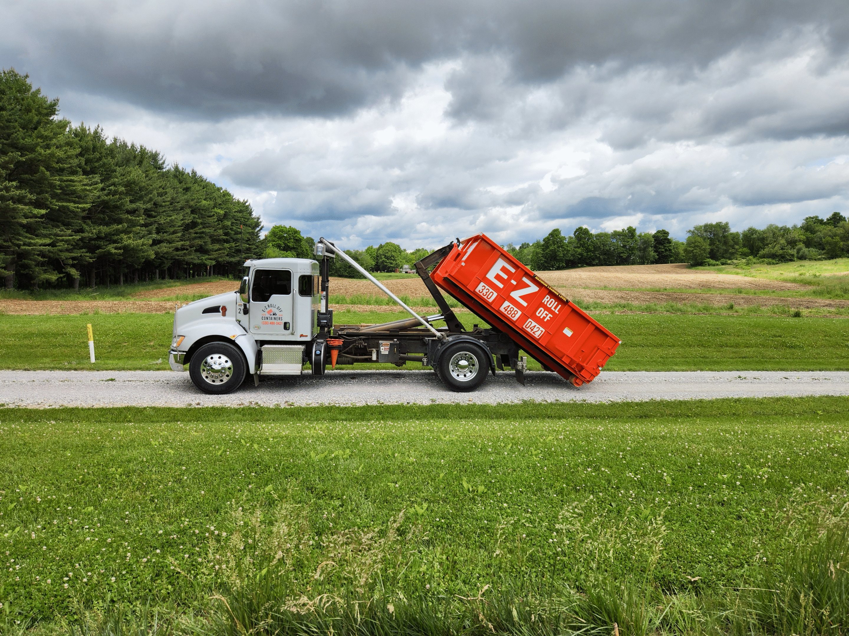 An orange roll-off dumpster placed in a residential driveway in Alliance, OH, beside a garage, surrounded by trees and a large yard on a sunny day.